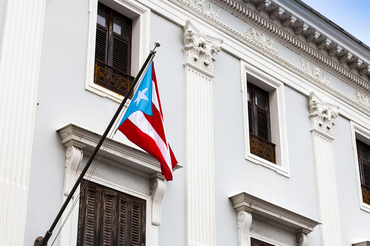 A Puerto Rican flag is mounted on a historic, white building with ornate architectural details and shuttered windows.