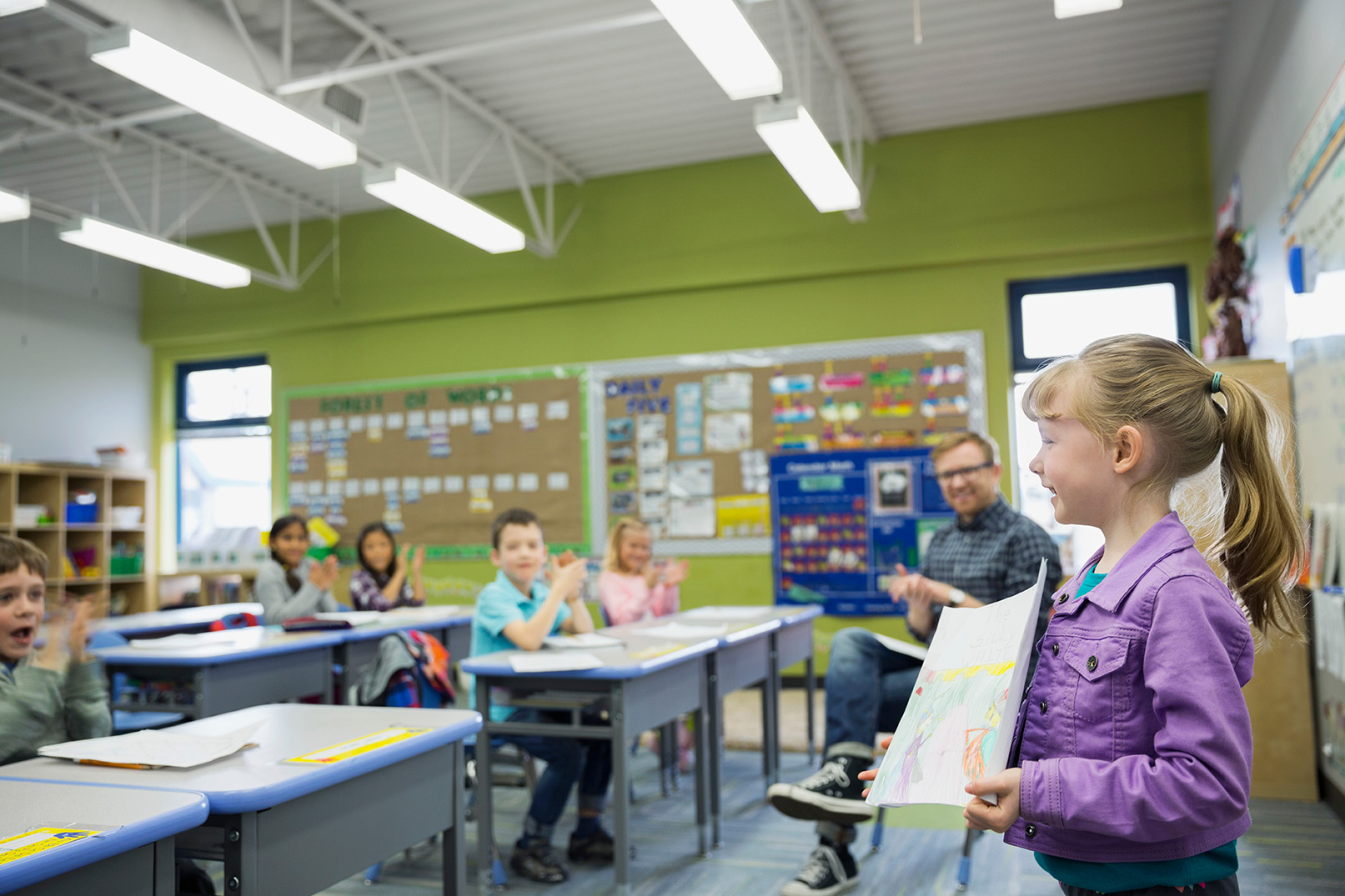 A young girl stands at the front of a classroom holding a drawing, while classmates and a teacher listen and watch from their desks.