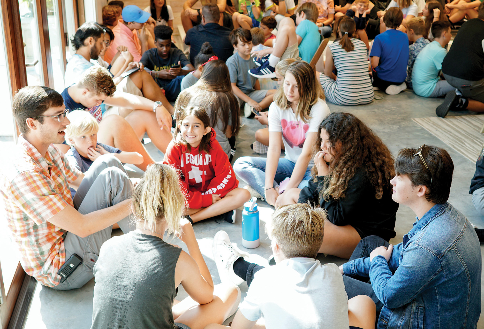 A large group of teens and a few adults sit closely together indoors, some talking in small circles, others listening or interacting with their phones.