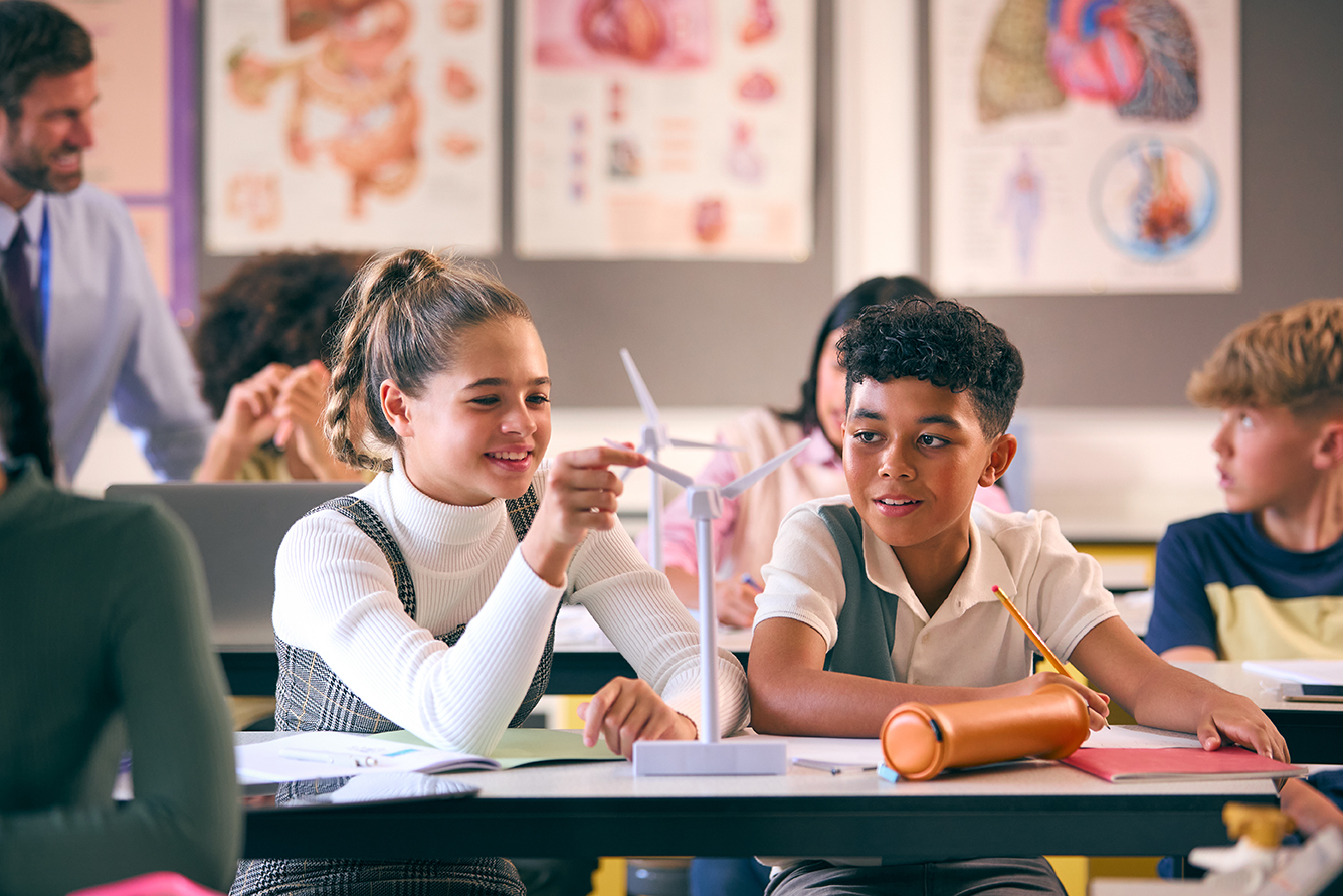Two students in a classroom examine a model wind turbine while other students and a teacher engage in background activities. Science diagrams are displayed on the walls.