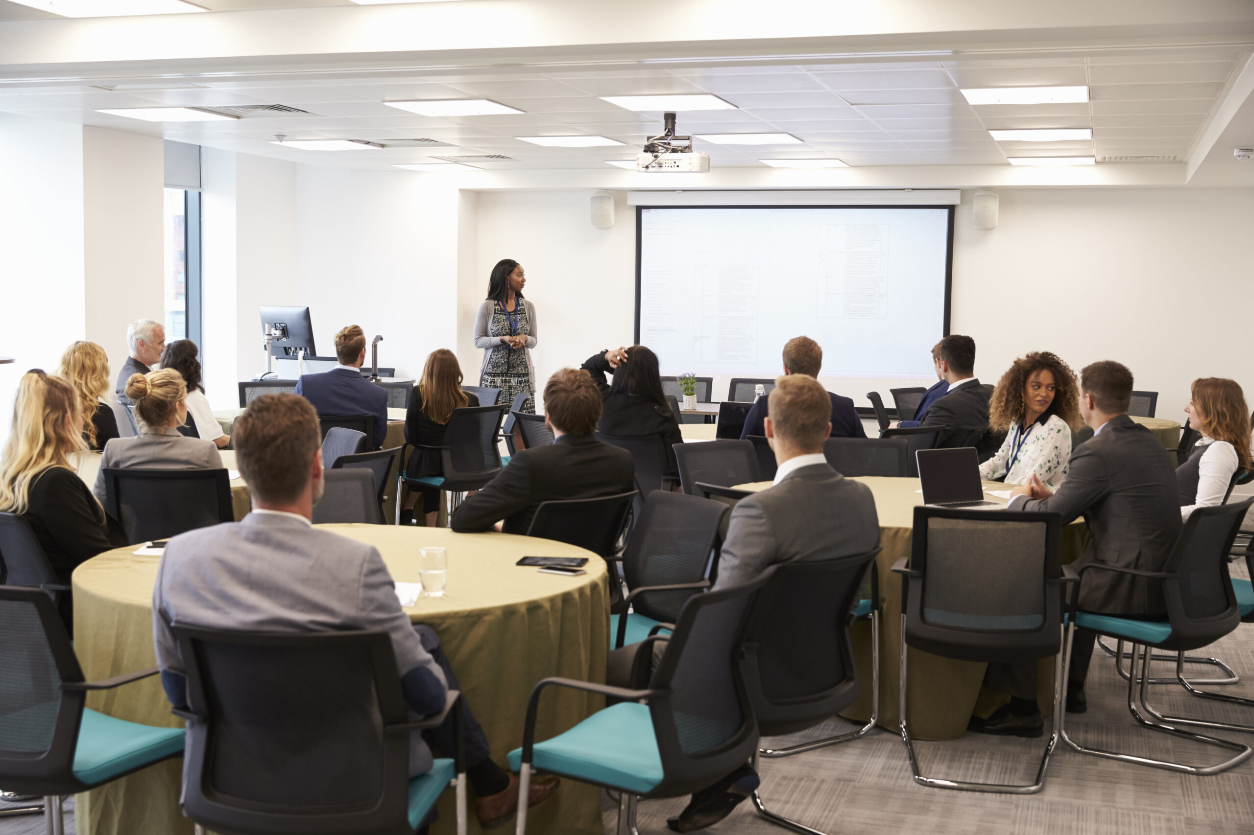 A group of people in business attire sit at round tables, listening to a woman presenting in front of a projection screen in a modern conference room.