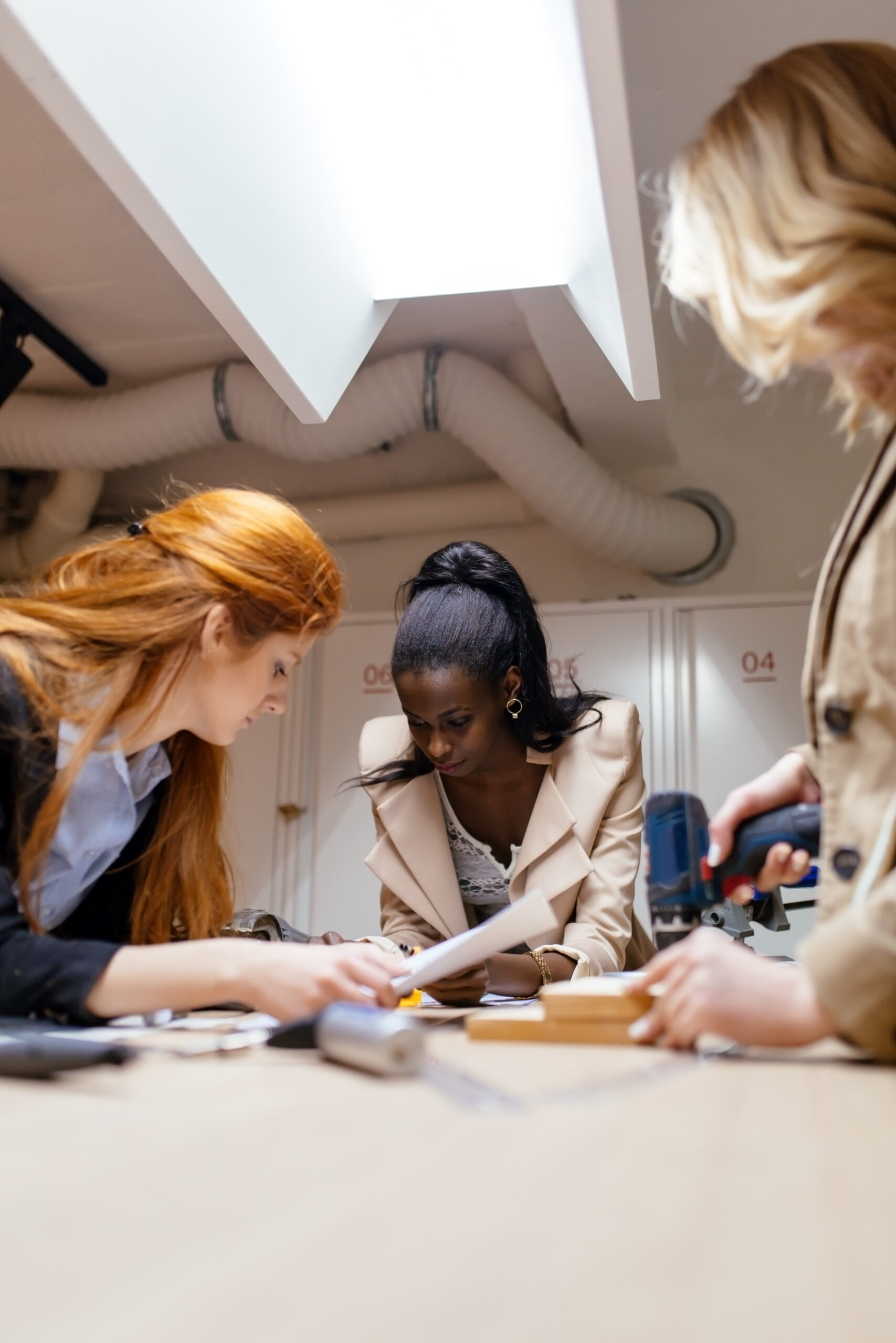 Three women collaborate on a project at a table, examining documents and using tools in a well-lit workspace.