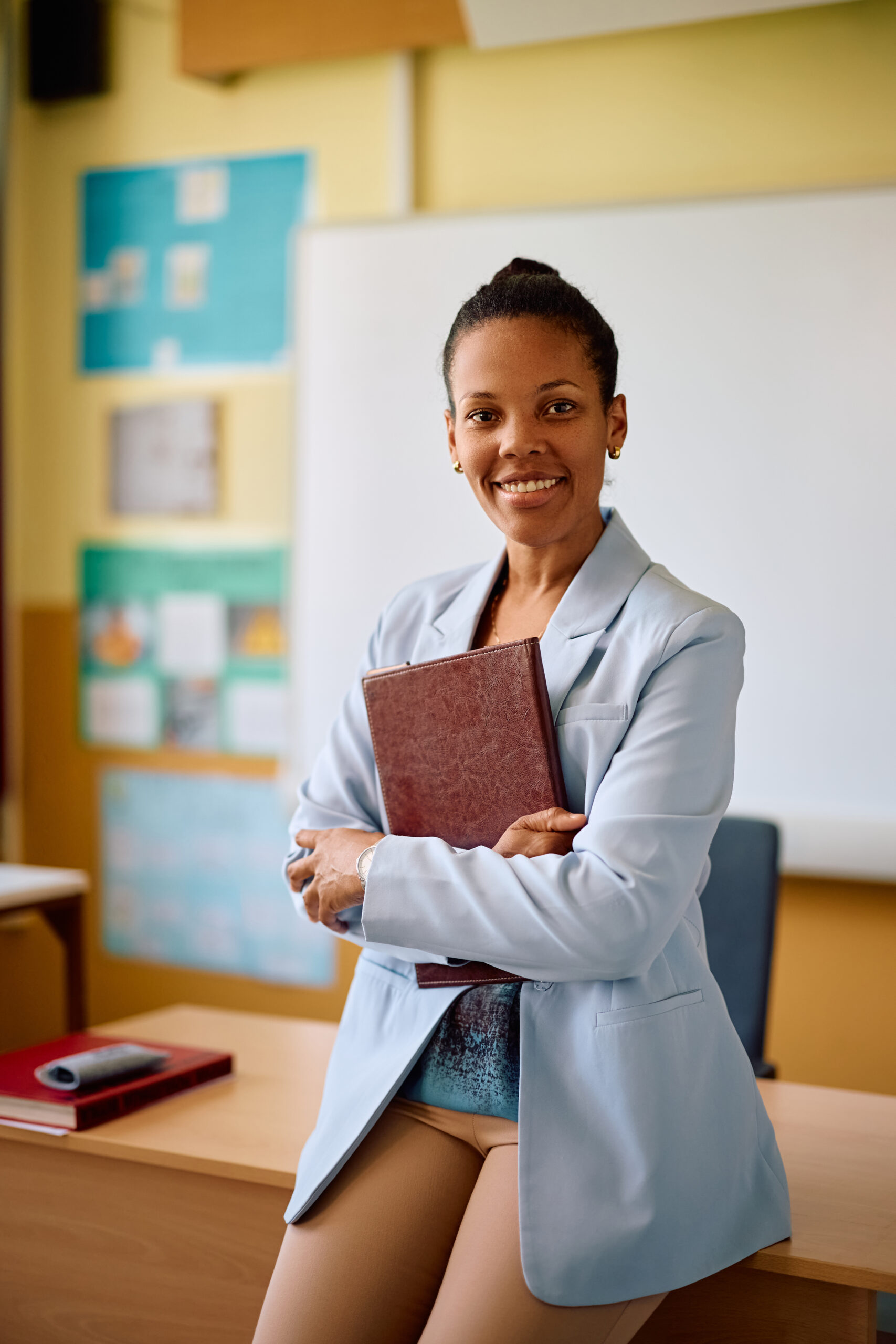 A woman in a light blue blazer stands in a classroom, holding a closed book and smiling, with a whiteboard and posters in the background.