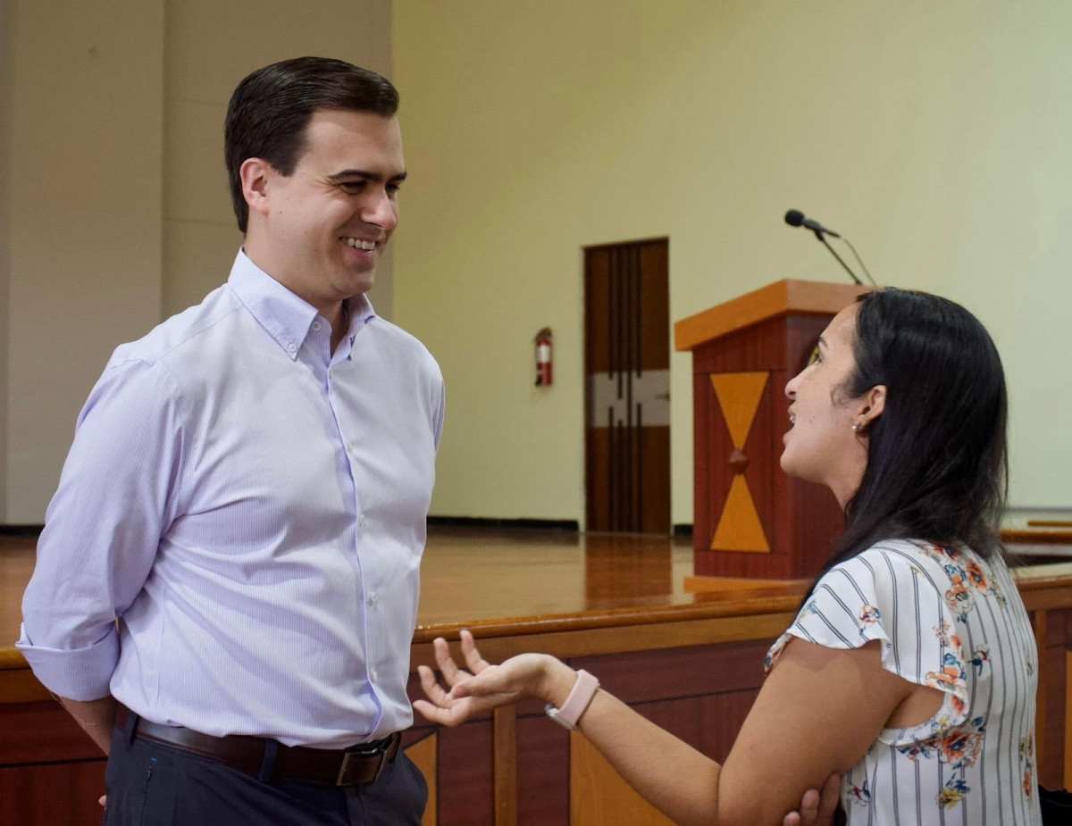 A man and a woman stand and talk in an auditorium near a wooden podium. The man is smiling and listening while the woman gestures with her hand.